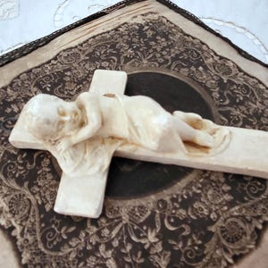 May include: A cream-coloured sculpture of a baby lying on a cross. The cross rests on a square, patterned fabric with a black lace border. The fabric has a dark brown and cream colour scheme.