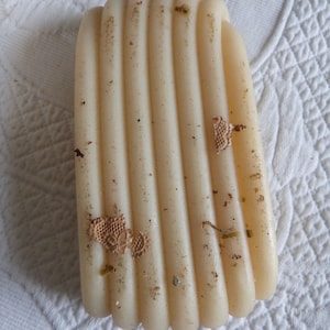 May include: A close-up of a beige, cylindrical candle with a textured surface. The candle is made of multiple layers of wax, creating a striped pattern. The candle has a few small, brown pieces of debris on its surface.