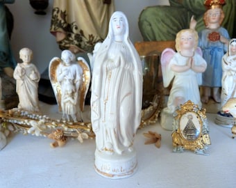 Puede incluir: Figurilla de porcelana blanca de la Virgen María, de pie con las manos juntas en oración. La figurilla lleva una larga túnica blanca con detalles dorados y una corona. La base de la figurilla está inscrita con las palabras "Niz ue to la".