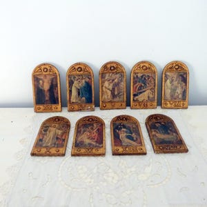 Könnte beinhalten: Eine Sammlung von zehn Vintage-Religionskunst-Tafeln. Jede Tafel zeigt eine detaillierte, farbenfrohe Illustration in einem dekorativen, gewölbten Holzrahmen. Römische Ziffern sind auf der Unterseite jeder Tafel zu sehen. Die Tafeln sind auf einer weißen Oberfläche angeordnet.