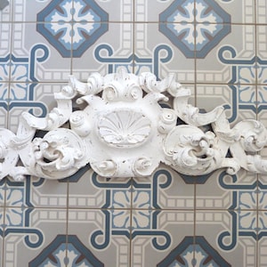 Puede incluir: Un elemento decorativo de pared blanco y ornamentado con un acabado envejecido. El elemento decorativo presenta un diseño central de concha y volutas, sobre un fondo de baldosas estampadas en tonos azules, grises y blancos. La pieza es probablemente de madera o material similar.