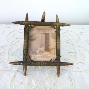 Könnte beinhalten: Ein Vintage-Bilderrahmen aus wiederverwendeten Patronenhülsen. Der Rahmen ist mit kleinen grünen Blättern verziert und hat eine rechteckige Öffnung für ein Foto. Der Rahmen steht auf einer weißen Spitzentischdecke.