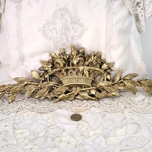 May include: A gold-colored decorative wall hanging featuring a basket overflowing with flowers and foliage. The ornate design includes roses and leaves, with a detailed basket weave pattern. The piece is set against a backdrop of white lace.