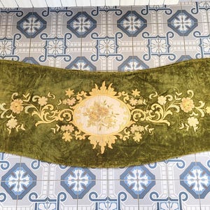Könnte beinhalten: Olivgrüner Samt-Volant mit gesticktem Blumendesign. Der Volant hat ein zentrales Oval mit einem Blumenstrauß und goldfarbenen Blumen- und Rankenverzierungen. Der Stoff hat ein weiches, strukturiertes Aussehen.