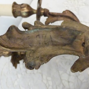 K&ouml;nnte beinhalten: Eine Wandleuchte aus Messing mit zwei Kerzenhaltern und verzierten Details. Die Leuchte hat eine dekorative R&uuml;ckwand mit einer Herstellermarke.
