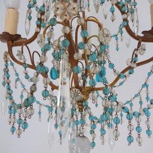 May include: Close-up of a vintage chandelier with a gold-colored frame. The chandelier is adorned with clear crystal pendants, turquoise and clear beads, and decorative floral accents. The light fixtures are visible at the top.