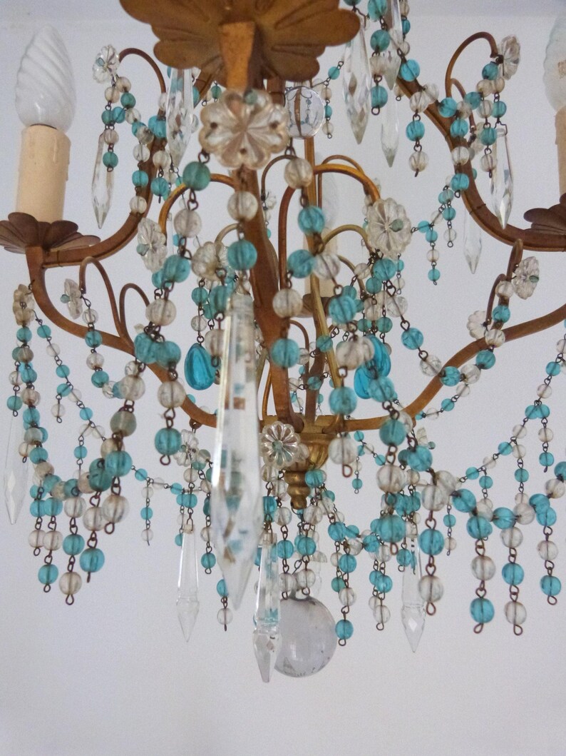 May include: Close-up of a vintage chandelier with a gold-colored frame. The chandelier is adorned with clear crystal pendants, turquoise and clear beads, and decorative floral accents. The light fixtures are visible at the top.