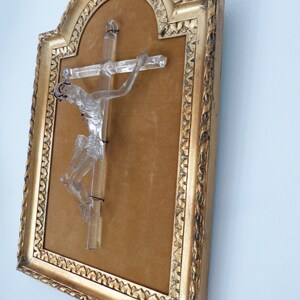 May include: A glass crucifix in a gold frame with a brown velvet backing. The crucifix is made of clear glass and depicts a figure of Jesus Christ.