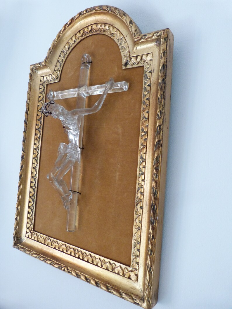 May include: A glass crucifix in a gold frame with a brown velvet backing. The crucifix is made of clear glass and depicts a figure of Jesus Christ.