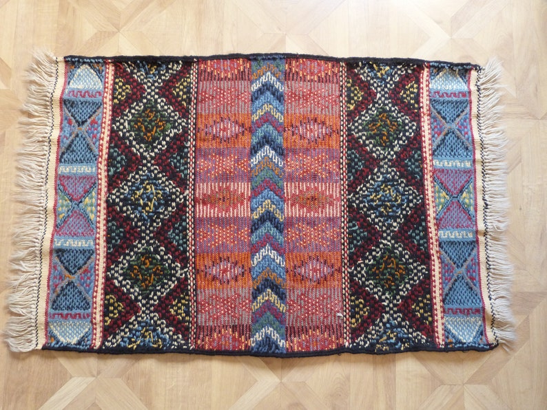 Op de afbeelding: Een kleurrijk geweven tapijt met een geometrisch patroon in tinten rood, blauw, zwart en wit. Het tapijt heeft aan alle kanten franjes.