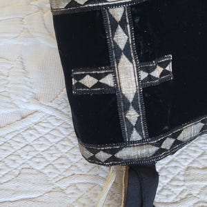 May include: Black velvet head covering with silver and diamond-patterned trim. A silver cross design is centered on the front. Cream-colored ties hang from the bottom. The item is displayed on a white, quilted surface.