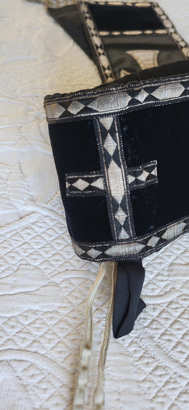 May include: Black velvet head covering with silver and diamond-patterned trim. A silver cross design is centered on the front. Cream-colored ties hang from the bottom. The item is displayed on a white, quilted surface.