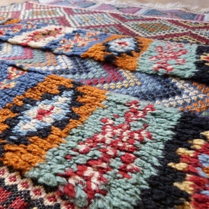 Op de afbeelding: Close-up van een kleurrijk geweven tapijt met ingewikkelde patronen in tinten blauw, rood, oranje en groen. Het tapijt heeft een textuur en is handgeweven.