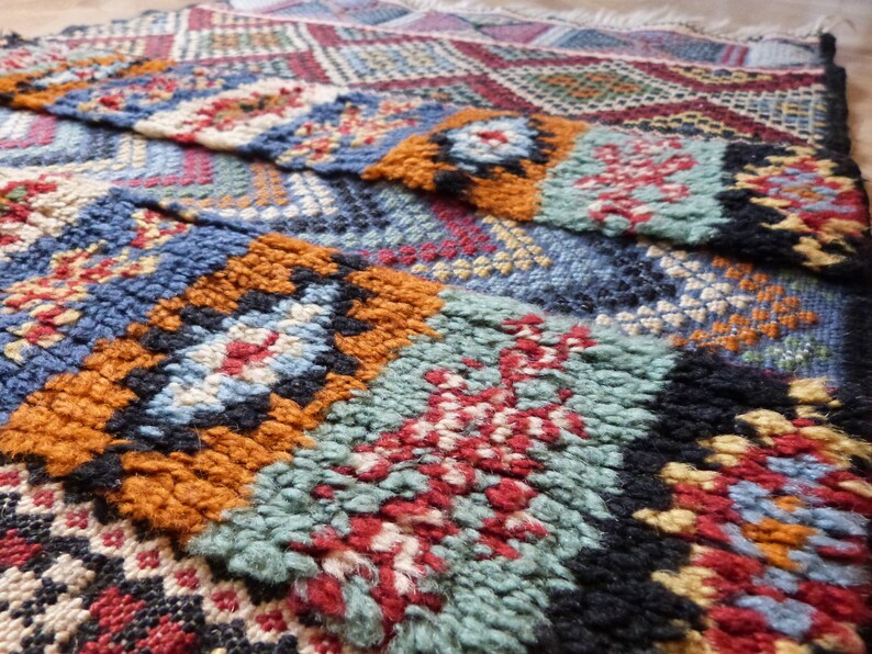 Op de afbeelding: Close-up van een kleurrijk geweven tapijt met ingewikkelde patronen in tinten blauw, rood, oranje en groen. Het tapijt heeft een textuur en is handgeweven.