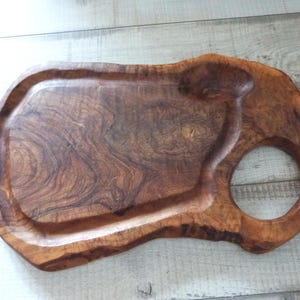 Könnte beinhalten: Ein Serviertablett aus Holz mit einer natürlichen, organischen Form. Das Tablett besteht aus dunkelbraunem Holz mit sichtbarer Maserung und einem kreisförmigen Griff. Es hat eine vertiefte Fläche.
