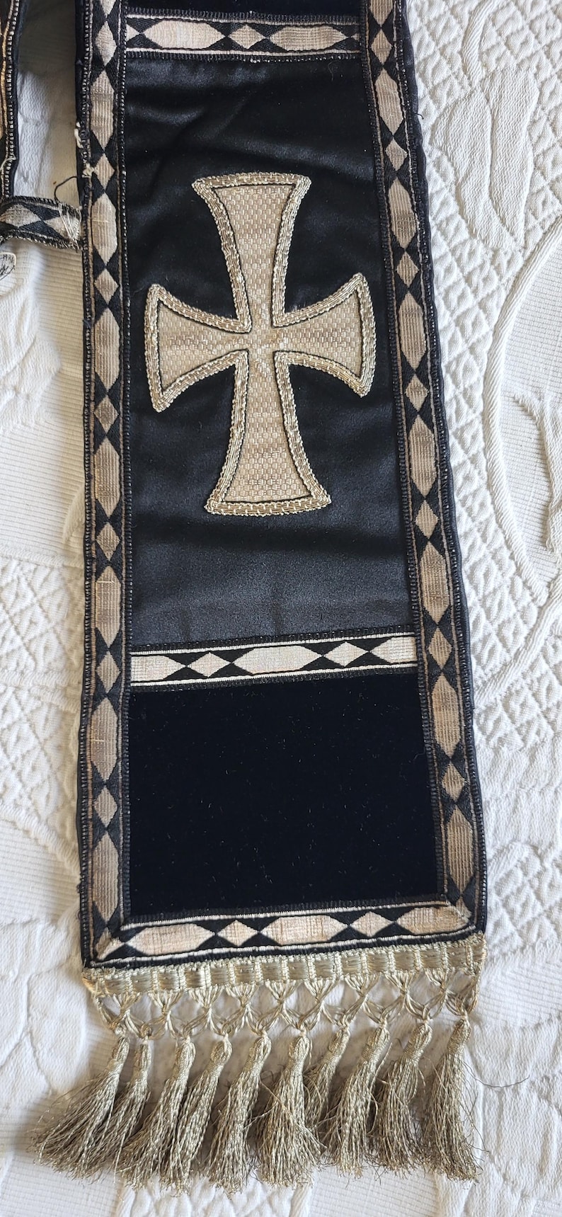 May include: Black and gold religious stole with a large cross design. The stole features a black velvet panel, gold trim, and a decorative fringe. The cross is embroidered with a gold-colored thread.