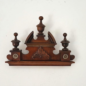 Può includere: Mensola in legno marrone scuro e ornata con una parte superiore decorativa. La mensola presenta tre pinnacoli, un motivo centrale intagliato e piccoli accenti circolari. Il pezzo è probabilmente vintage o antico.