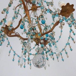 May include: An antique chandelier with a bronze-colored frame, adorned with clear and turquoise-colored beads and crystal pendants. The fixture has two light bulbs and a decorative glass globe at the bottom, creating a vintage aesthetic.