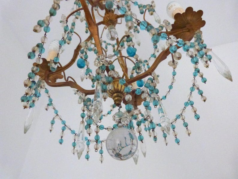 May include: An antique chandelier with a bronze-colored frame, adorned with clear and turquoise-colored beads and crystal pendants. The fixture has two light bulbs and a decorative glass globe at the bottom, creating a vintage aesthetic.
