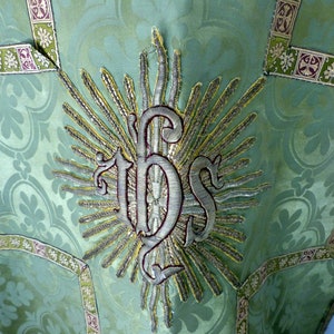 May include: Close-up of a green liturgical vestment with a floral pattern. The vestment features an embroidered IHS symbol in silver and gold, surrounded by rays. Decorative trim in green, gold, and purple accents the edges.