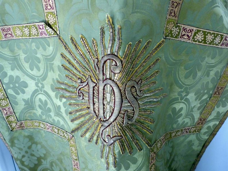 May include: Close-up of a green liturgical vestment with a floral pattern. The vestment features an embroidered IHS symbol in silver and gold, surrounded by rays. Decorative trim in green, gold, and purple accents the edges.
