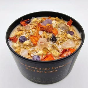 May include: A black candle jar filled with a creamy wax, adorned with crystals, dried flowers, and herbs. The candle is labeled "Energy Clearing and Recharging Spell Jar Candle" and is designed for spiritual use.