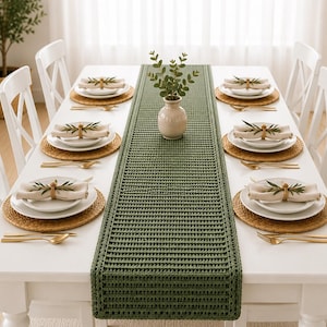 May include: A long, rectangular, olive green crocheted table runner with a textured, ribbed stitch pattern. The runner is centred on a white dining table set with six white plates, woven placemats, gold cutlery, and linen napkins with greenery.