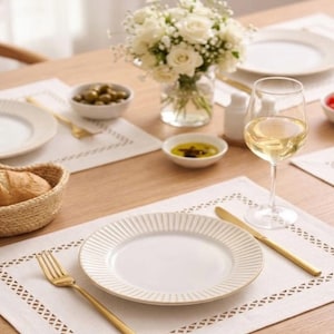 May include: A beautifully set table with white plates, gold cutlery, and a glass of white wine. A small bouquet of white roses sits in a glass vase. The table setting includes olives, bread, and olive oil.
