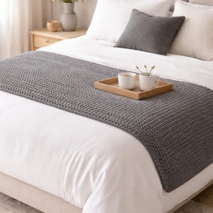 Pode incluir: Um corredor de cama cinza tricotado é colocado sobre uma cama branca. Uma bandeja de madeira com duas canecas brancas está sobre o corredor de cama. A cama é feita com lençóis e travesseiros brancos, com uma almofada cinza e um vaso de flores.