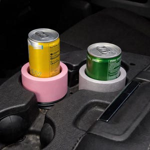 Puede incluir: Dos insertos para portavasos de coche, uno rosa y otro gris, cada uno con una lata de bebida. El inserto rosa contiene una lata amarilla y el inserto gris una lata verde. Los portavasos están en la consola central de un coche.