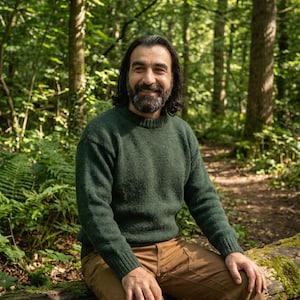 May include: A man wearing a green knit sweater and brown pants sits on a log in a forest. The sweater has a crew neck and ribbed cuffs. The man has long dark hair and a beard, smiling at the camera.