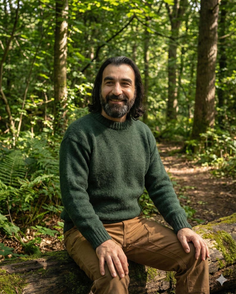 May include: A man wearing a green knit sweater and brown pants sits on a log in a forest. The sweater has a crew neck and ribbed cuffs. The man has long dark hair and a beard, smiling at the camera.