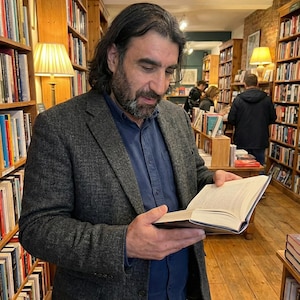 May include: A man in a gray blazer and blue shirt reads a book in a bookstore. Bookshelves filled with books line the walls. The scene is illuminated by warm lighting from lamps and overhead fixtures.
