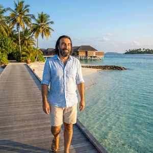 May include: A man wearing a light blue button-down shirt, khaki shorts, and brown sandals walks on a wooden boardwalk. The boardwalk leads to a beach with palm trees and overwater bungalows. The water is clear and turquoise.