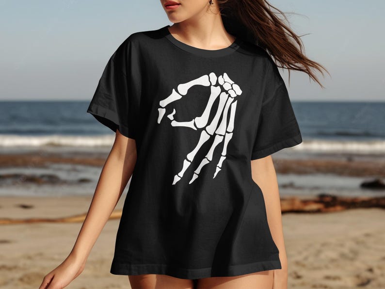 Black t-shirt with a white skeleton hand gesture graphic on a beach.