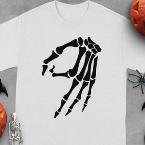 Black skeleton hand forming the "okay" gesture on a white t-shirt.