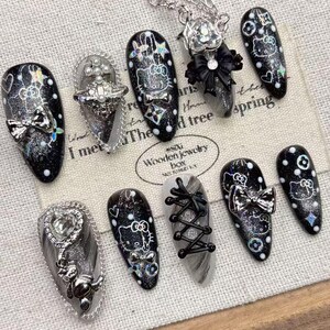 Può includere: Un set di dieci unghie finte con base nera glitterata, contorni bianchi del personaggio Hello Kitty, fiocchi metallici argentati, strass, una catenella argentata e dettagli a lacci tipo corsetto neri su un tessuto bianco sporco testurizzato.