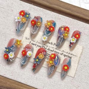 Può includere: Un set di dieci unghie finte lunghe a forma di mandorla. Ogni unghia presenta un design unico con decorazioni floreali colorate in rosso, giallo, blu e bianco. Alcune unghie hanno accenti iridescenti e piccoli strass. Le unghie sono disposte su una superficie chiara.