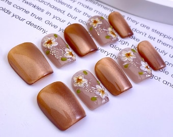 May include: A set of ten press-on nails featuring a mix of solid metallic bronze cat-eye polish and translucent nude nails adorned with hand-painted white flowers, green leaves, and silver glitter accents arranged on a white paper background.