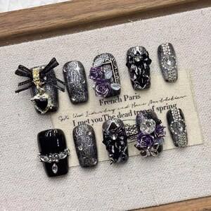 Può includere: Un set di dieci unghie finte a tema gotico. Le unghie sono nere, argentate e viola, con abbellimenti tra cui fiocchi, strass e disegni floreali. Il testo "French Paris" è visibile.