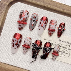 Può includere: Una collezione di unghie finte con vari design. Alcune unghie presentano gocce di glitter rosso, motivi floreali o una croce con gemme rosse. Altre hanno accenti di ali di pipistrello e catene. Le unghie sono esposte su una superficie chiara.