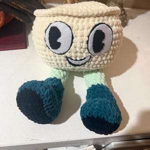 May include: A handmade crochet creature with a cream-colored head, large black and white eyes, and a smiling mouth. The legs are green and teal, with dark teal feet. The plush toy is sitting on a white surface.