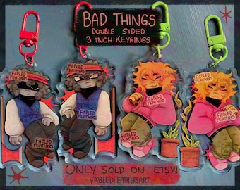 May include: Four double-sided 3-inch acrylic keyrings featuring illustrated characters with colorful clothing and accessories. Two keyrings have red clips and two have lime green clips. Text on the display reads BAD THINGS, DOUBLE SIDED, 3 INCH KEYRINGS, ONLY SOLD ON ETSY, and FABLEDFEATHERSART.