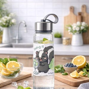 Può includere: Una bottiglia d'acqua in vetro trasparente con coperchio in acciaio inossidabile e manico grigio. La bottiglia presenta una grafica di puzzola dei cartoni animati con la scritta "fixing it". La bottiglia è riempita con acqua, fette di limone e foglie di menta.