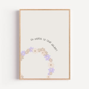 Könnte beinhalten: Gerahmter Druck mit hellbeigem Hintergrund. Der Druck zeigt einen Blumenkranz in Lila-, Beige- und Weißtönen am unteren Rand. Über dem Kranz steht der Text "Ich werde so sehr geliebt" in Handschrift.