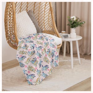 Puede incluir: Una manta rosa claro con un estampado repetido de perros de dibujos animados y flores rosas est&aacute; sobre una silla de mimbre. Una almohada blanca y una gris est&aacute;n dentro de la silla. Una peque&ntilde;a mesa blanca con un jarr&oacute;n de flores est&aacute; al lado de la silla.