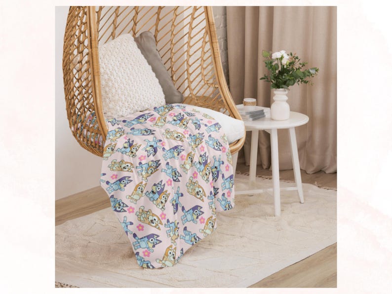Puede incluir: Una manta rosa claro con un estampado repetido de perros de dibujos animados y flores rosas est&aacute; sobre una silla de mimbre. Una almohada blanca y una gris est&aacute;n dentro de la silla. Una peque&ntilde;a mesa blanca con un jarr&oacute;n de flores est&aacute; al lado de la silla.