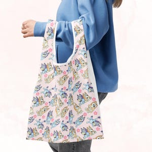 Puede incluir: Un bolso tote rosa con un patr&oacute;n repetido de perros de dibujos animados en varias poses, con detalles en azul, beige y blanco. El bolso tiene un interior blanco y un asa blanca.
