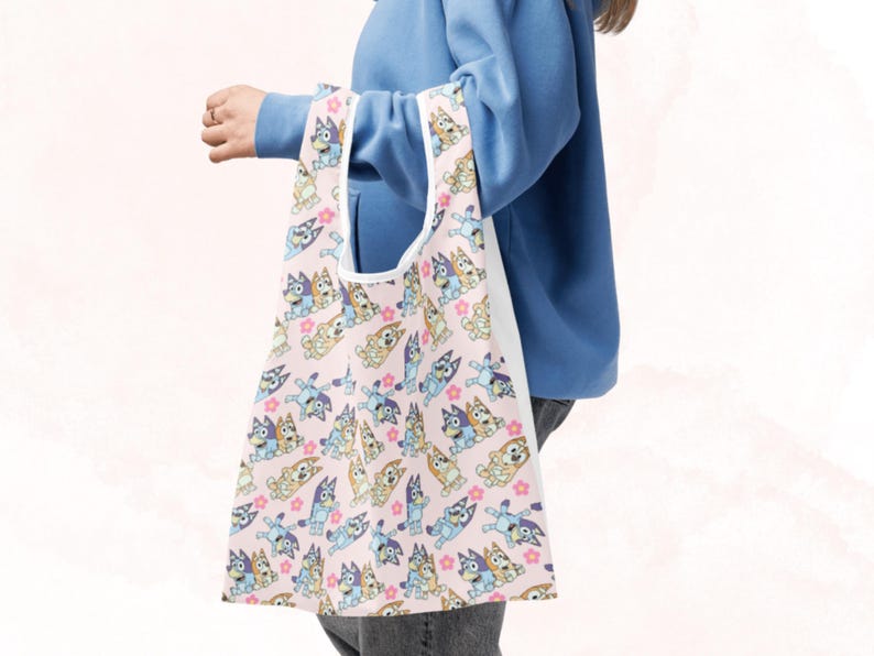 Puede incluir: Un bolso tote rosa con un patr&oacute;n repetido de perros de dibujos animados en varias poses, con detalles en azul, beige y blanco. El bolso tiene un interior blanco y un asa blanca.