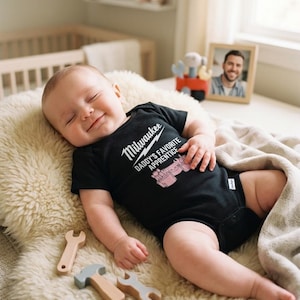 May include: A smiling baby wearing a black onesie with the text "Milwaukee Daddy's Favorite Apprentice" lies on a fluffy, cream-colored blanket. Wooden toy tools are scattered nearby. A framed photo and a crib are in the background.